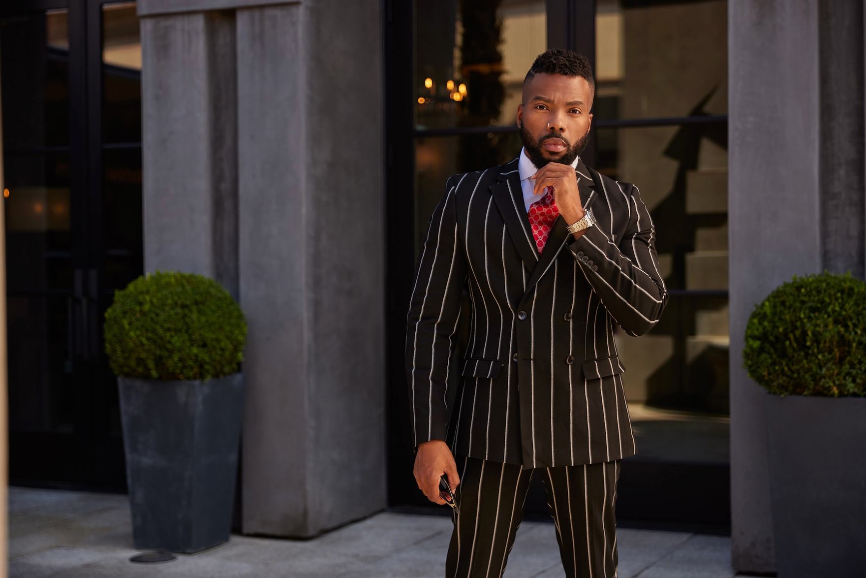 Sike Akhionbare standing in a chalk-stripe double-breasted suit, hand at his chin